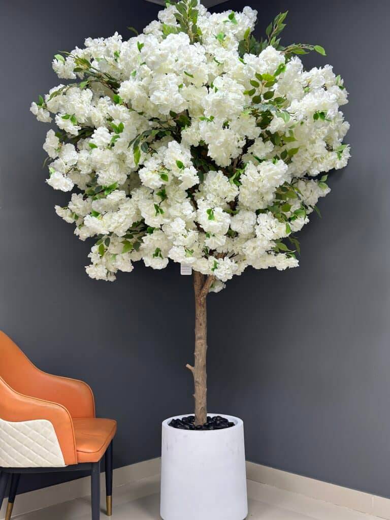 Large artificial white cherry blossom tree with a natural wood trunk in a white minimalist pot against a dark grey wall.